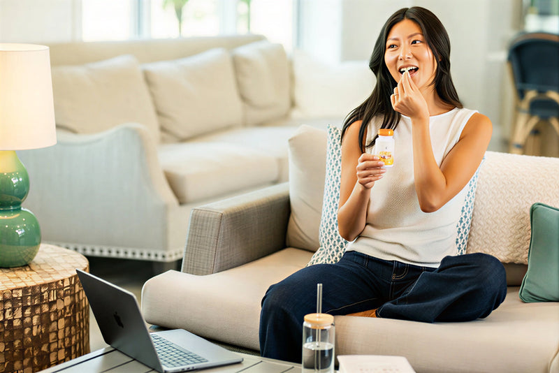 A woman sits on a beige sofa in a bright living room, holding a small pill bottle and bringing a pill to her mouth. A laptop is on the table in the foreground, with a lamp and another sofa in the background.