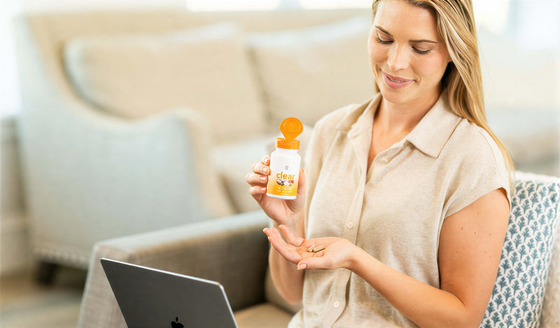 A woman sitting in a living room holds a bottle of Clear liquid supplements and pours some into her hand, with a laptop in the foreground and a sofa behind her.