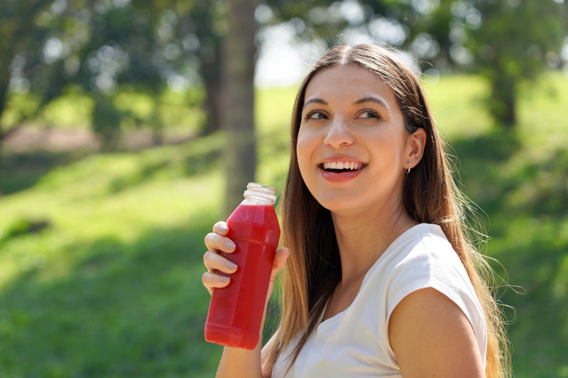 A woman smiling outdoors while holding a clear plastic bottle filled with red juice. She is wearing a white t-shirt, and the background shows a blurred green grassy field with trees.