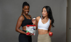 Two women standing indoors against a gray wall, one holding a red and white box labeled 'Gochujang' and the other holding a small jar. Both are dressed in athletic wear, and a red apple is visible in the hand of the woman on the right.