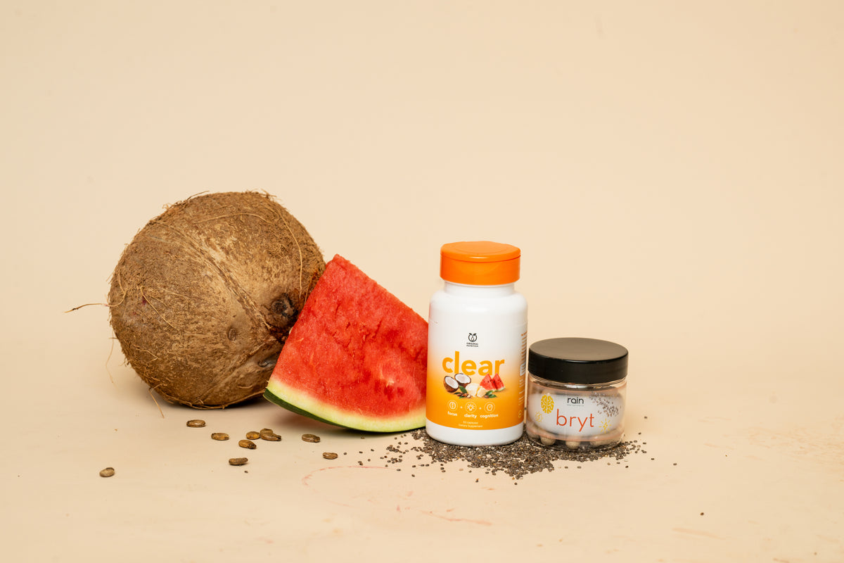 A supplement product display featuring a white bottle labeled 'clear' with an orange cap, a small jar labeled 'bryl' with a black lid, and fresh ingredients including a whole coconut, a slice of watermelon, and seeds on a beige background.