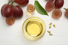 A glass bowl filled with golden yellow oil, surrounded by red grapes, sliced grapes, a green leaf, and a small jar on a white wooden background.