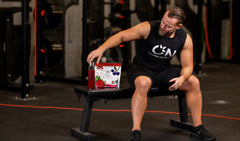 A man in a gym setting wearing a black sleeveless shirt with a white 'C&N' logo sits on a weight bench, reaching toward a red and white bottle on the bench.