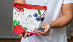 A person in a white shirt holds a bag of ON Flow red berry supplement powder in one hand and a small handful of blueberries in the other, against a neutral indoor background.
