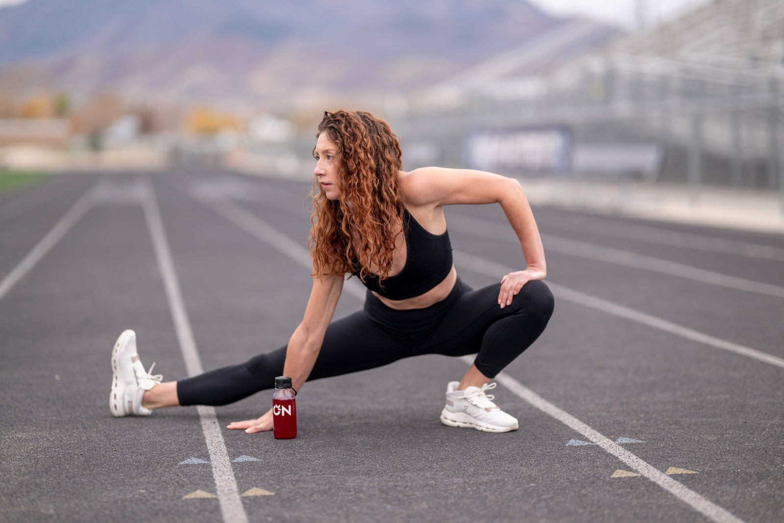 A woman in a black sports bra and leggings stretches on a running track, with a small red bottle labeled CNN near her foot. The background shows a blurred outdoor stadium and mountains.