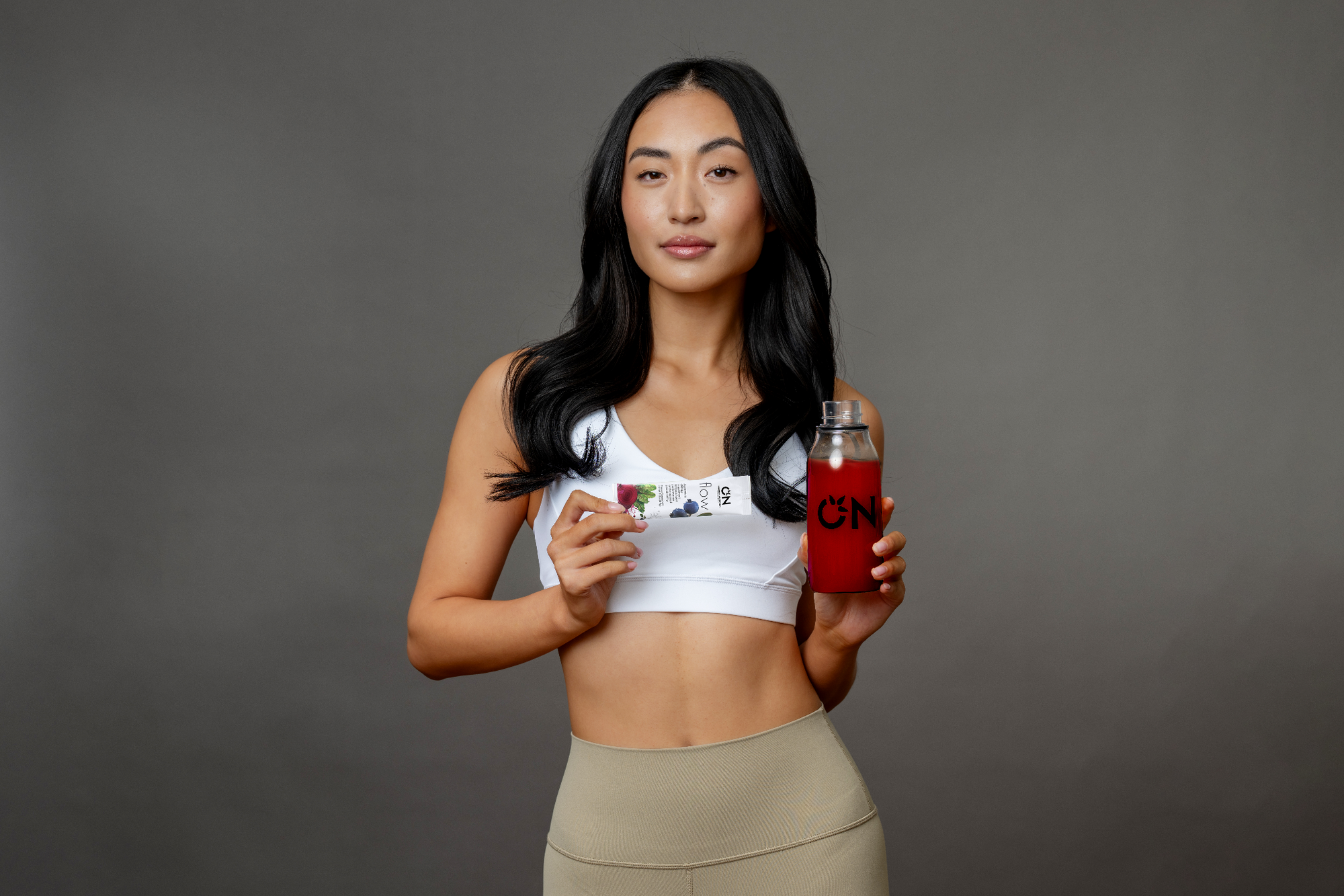 A woman in athletic wear holding a supplement packet and a bottle filled with red liquid.