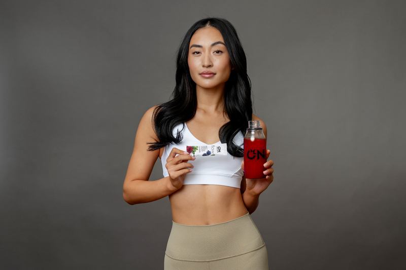 A woman in athletic wear holding a supplement packet and a bottle filled with red liquid.