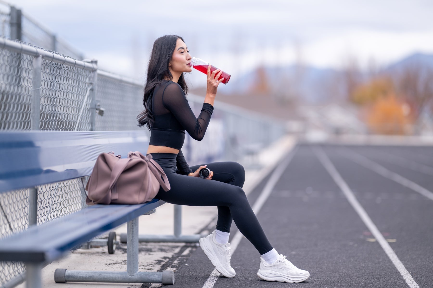 A woman in black athletic wear sitting on a bench at a track and drinking a red liquid from a bottle.
