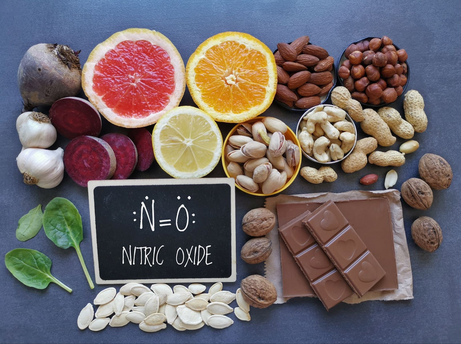 Assorted foods including fruits, nuts, and chocolate with a chalkboard sign displaying 'Nitric Oxide' on a blue background.