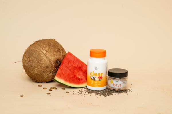 A supplement product display featuring a white bottle labeled 'clear' with an orange cap, a small jar labeled 'bryl' with a black lid, and fresh ingredients including a whole coconut, a slice of watermelon, and seeds on a beige background.